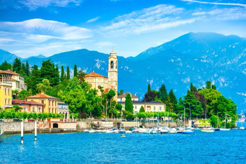 A church bell tower, villas and sailboats on the shore of Lake Como, with mountains behind